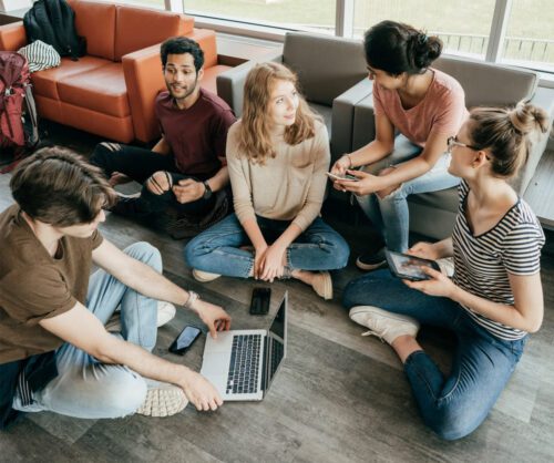 Cinco jóvenes sentados en el suelo y sillones de una sala común, conversando mientras usan un portátil, una tablet y teléfonos móviles.