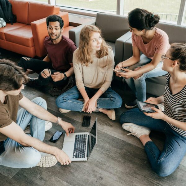 Cinco jóvenes sentados en el suelo y sillones de una sala común, conversando mientras usan un portátil, una tablet y teléfonos móviles.